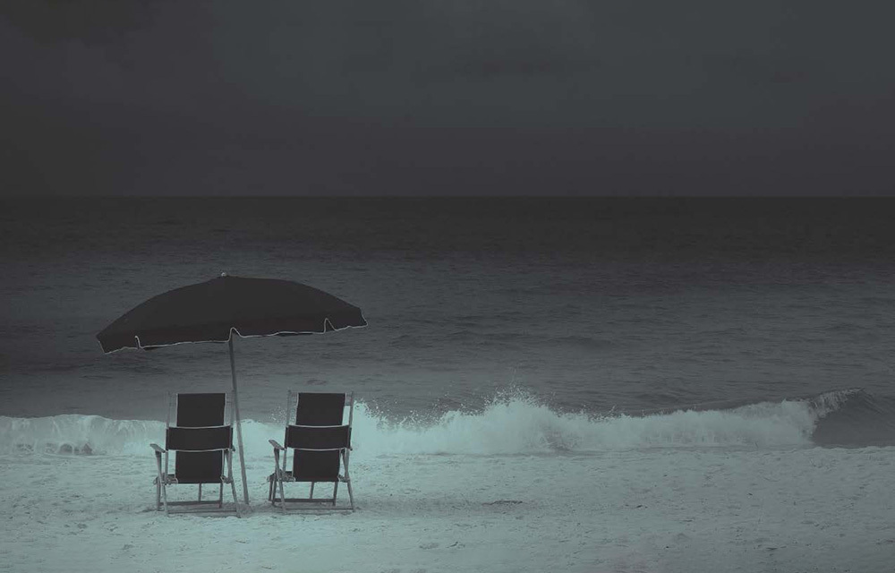 Two beach chairs beside each other as small waves crash onto the beach. The scene is filtered with dark, gloomy colors.