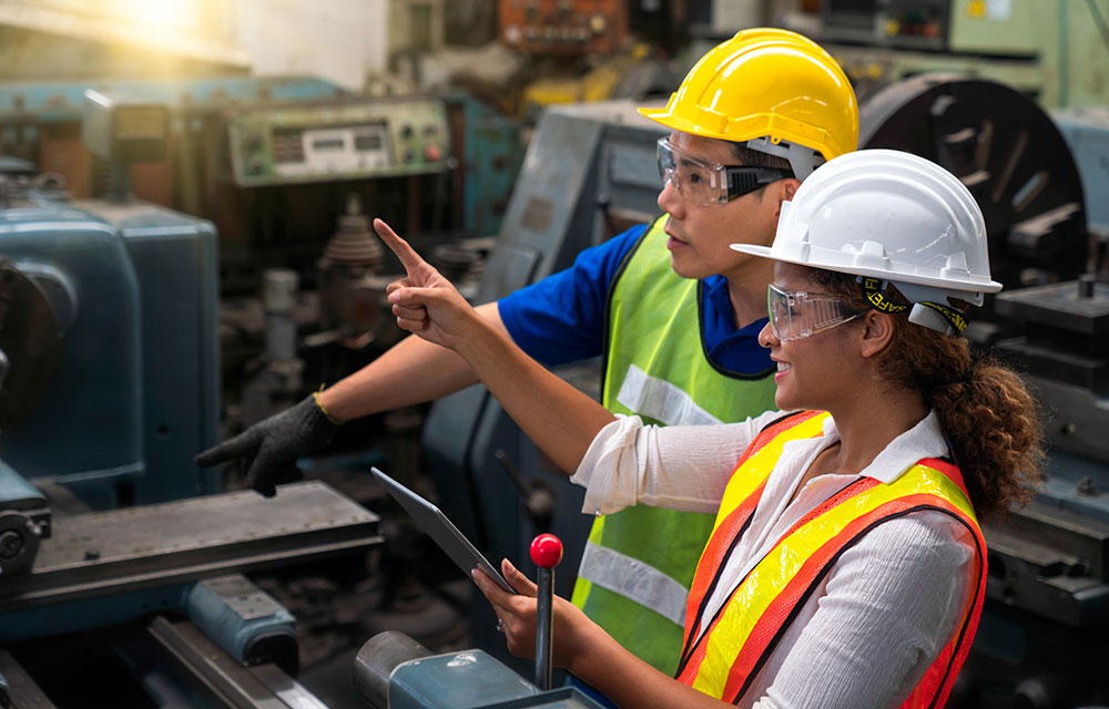 Two factory workers in hard hats and safety vests review the guide for a piece of equipment.