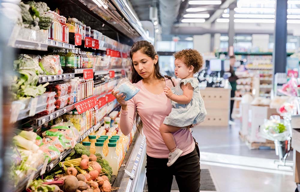 Woman holds a toddler while examining groceries at a grocery store