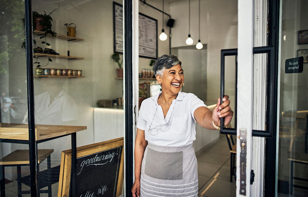Smiling business owner stands holding the door open to their small business