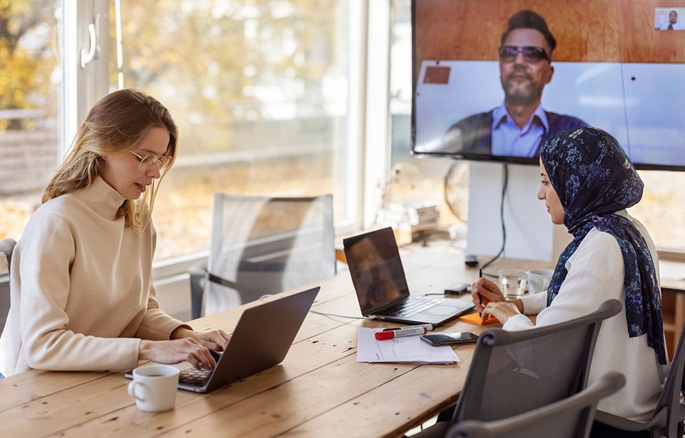 Two workers sitting at a table with laptops in front of them for virtual meeting with an at home worker on a larger screen hanging on the wall.