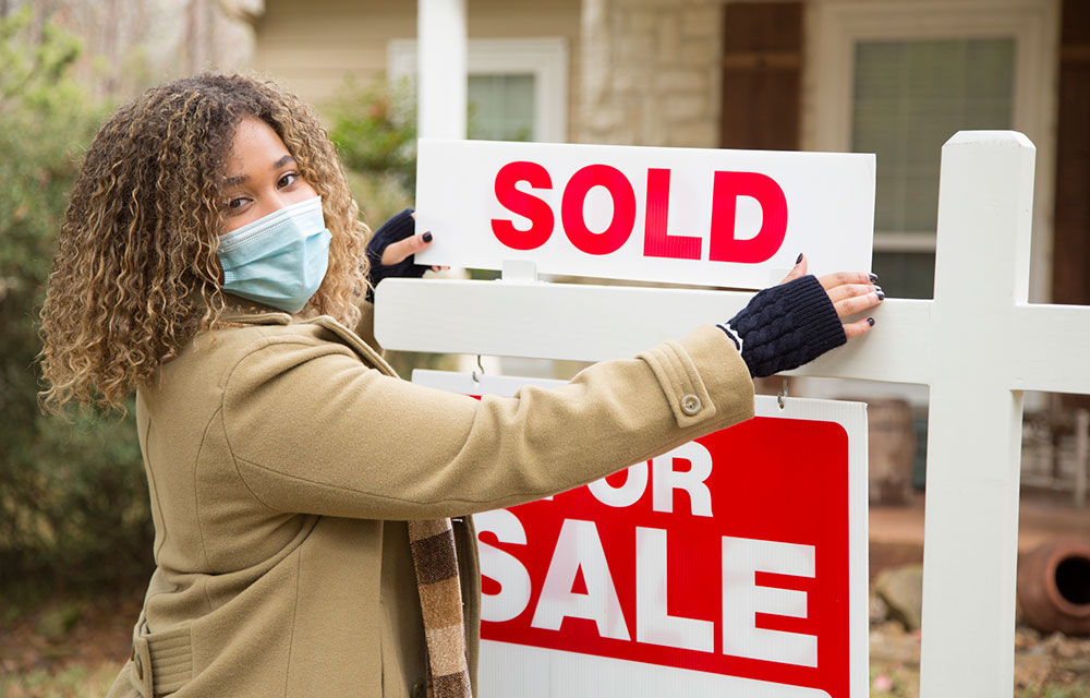 masked woman adding a sold sign to a for sale house sign