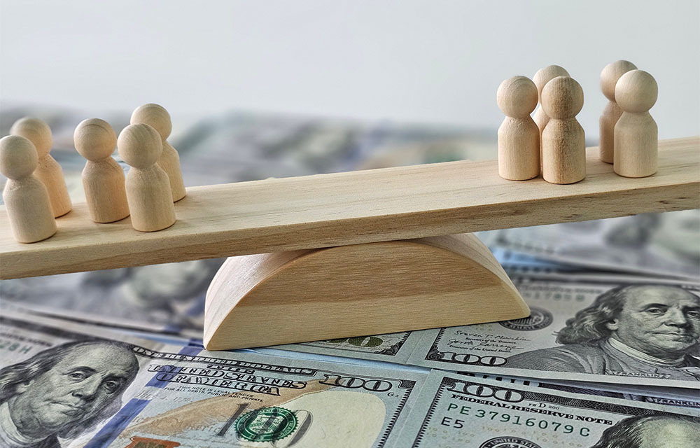A teetering seesaw with wooden figurines representing people balanced on each side sits on a pile of money.