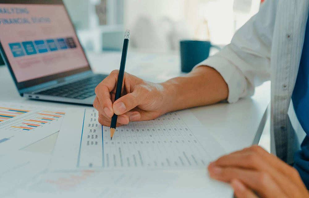 Close up of a person making pencil marks on a printed-out chart with a spread of reports surrounding.