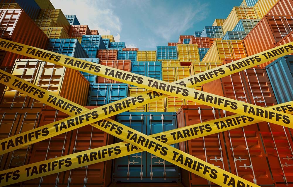 Wall of shipping containers with caution tape labeled with "tariffs" stretched across.