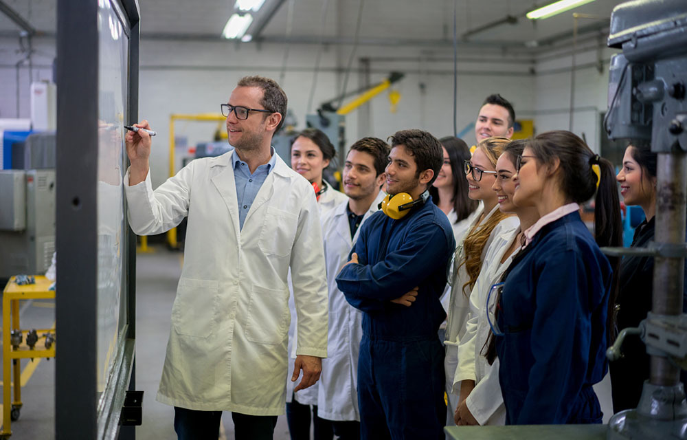 Students in blue overalls smiling and intrigued as their professor writes on a whiteboard.