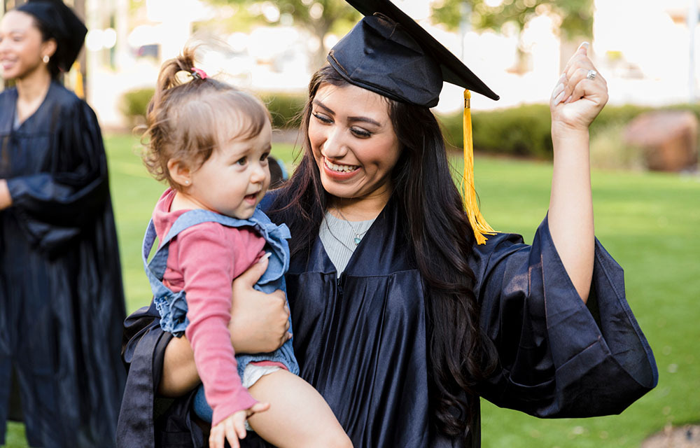 Young adult mother, in her cap and gown, smiles as she teaches her baby daughter how to cheer.
