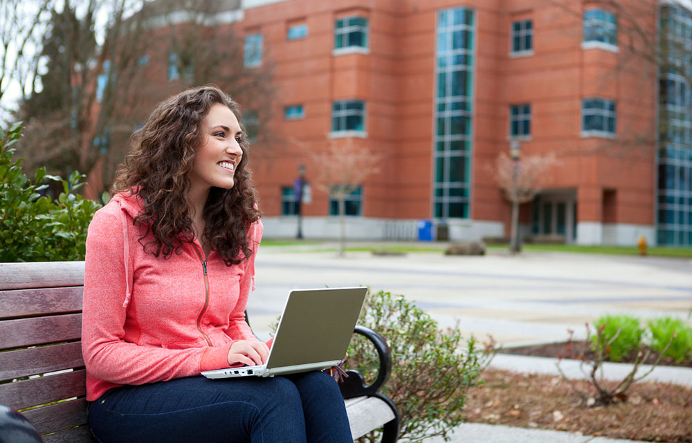 A happy young woman sits outside of a community college while using her laptop.