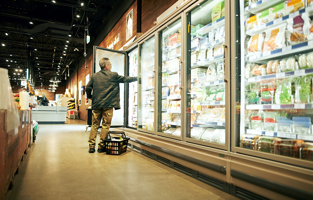 Man looking at prices and grabbing food from the freezer section of a grocery store.