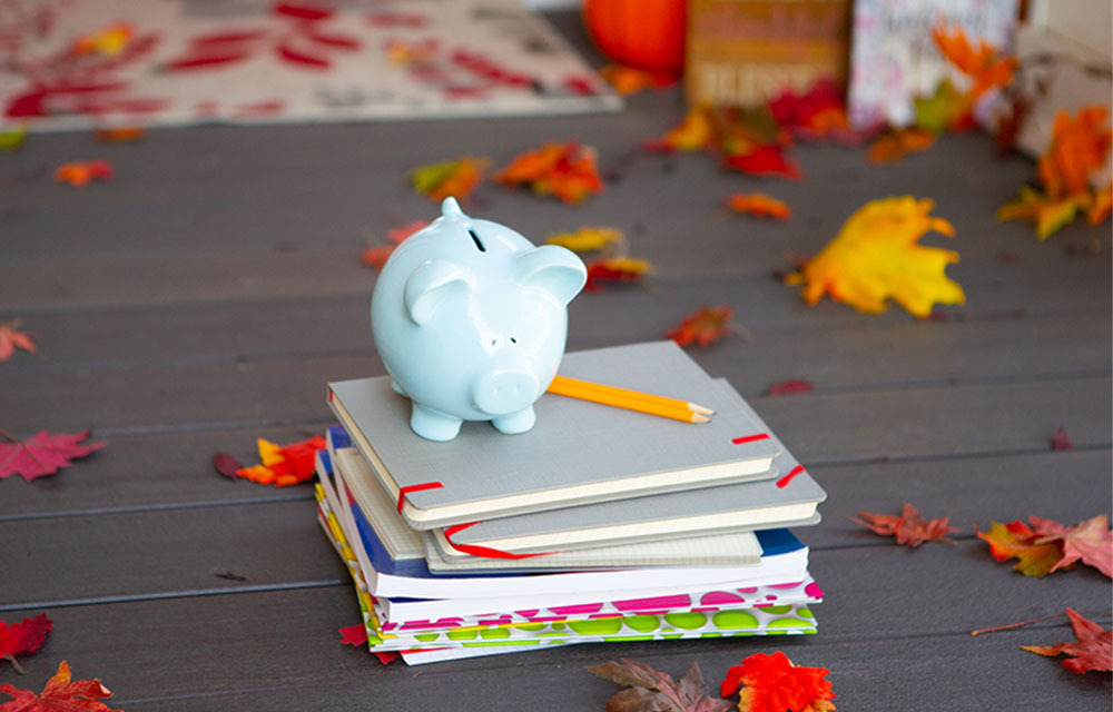 Blue piggy bank on top of a stack of notebooks laying on a porch sprinkled with Fall leaves.