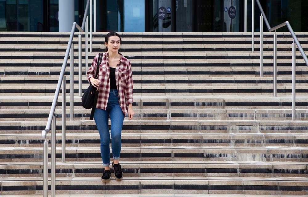 Student wearing a checkered and jeans and carrying a shoulder bag walks down the stairs from their college campus.