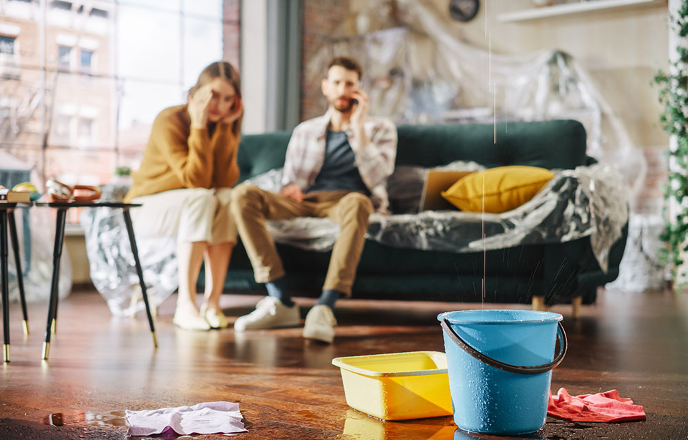 A leaky roof is dripping into a bucket with a stressed out couple in the background.