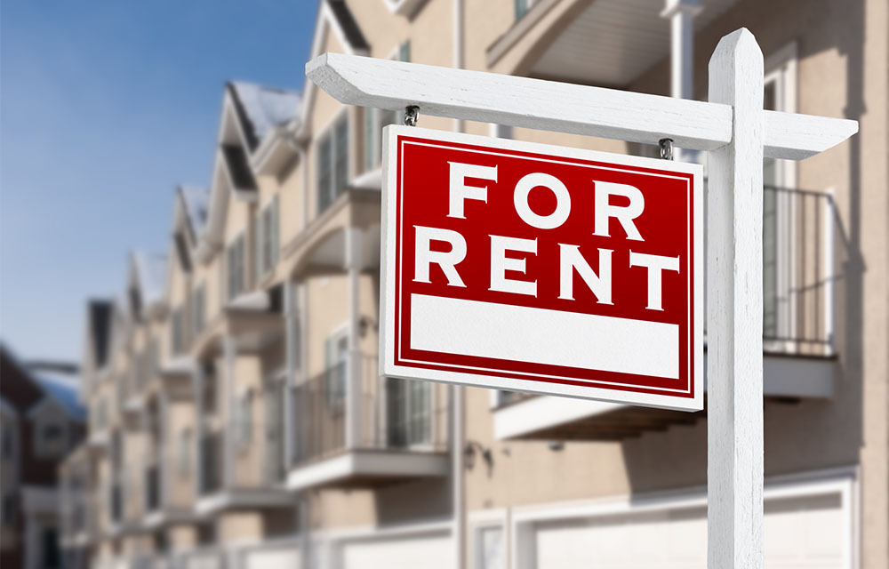 For rent sign in front of a row of apartments with balconies and garage doors.
