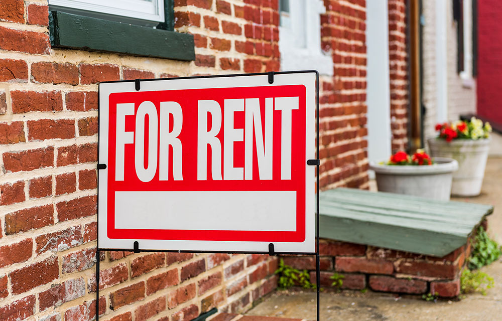 Red for rent sign stuck in the ground in front of brick house.