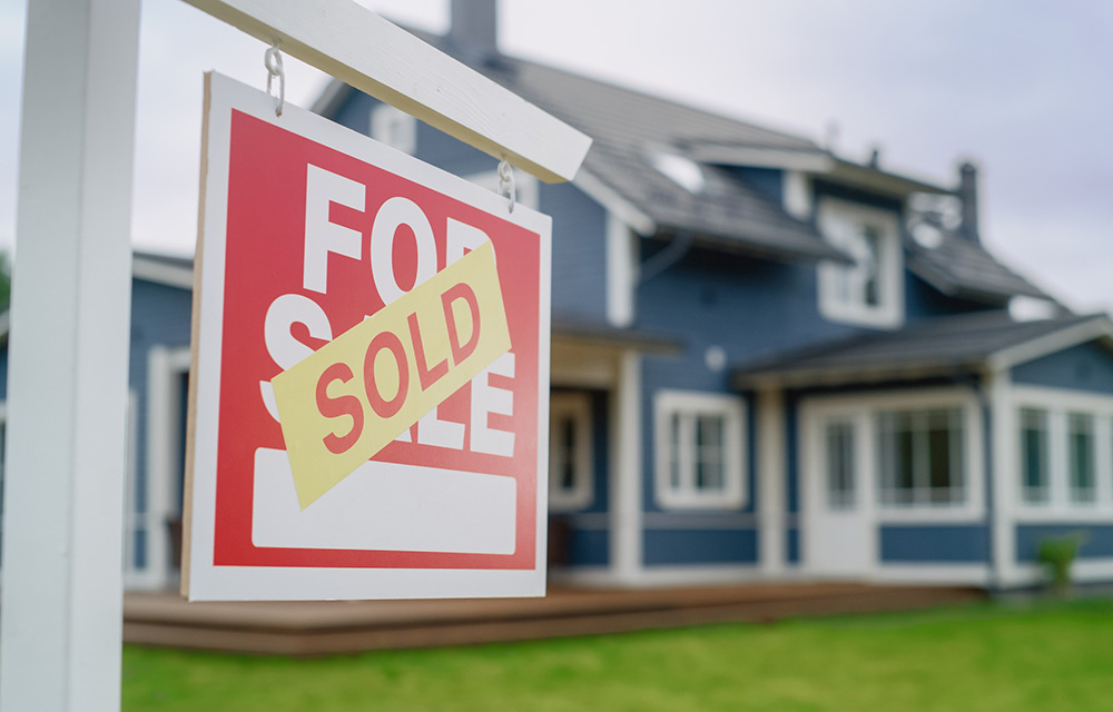 For sale sign hanging on post with sold sticker stuck on the front and a medium sized house in the background.