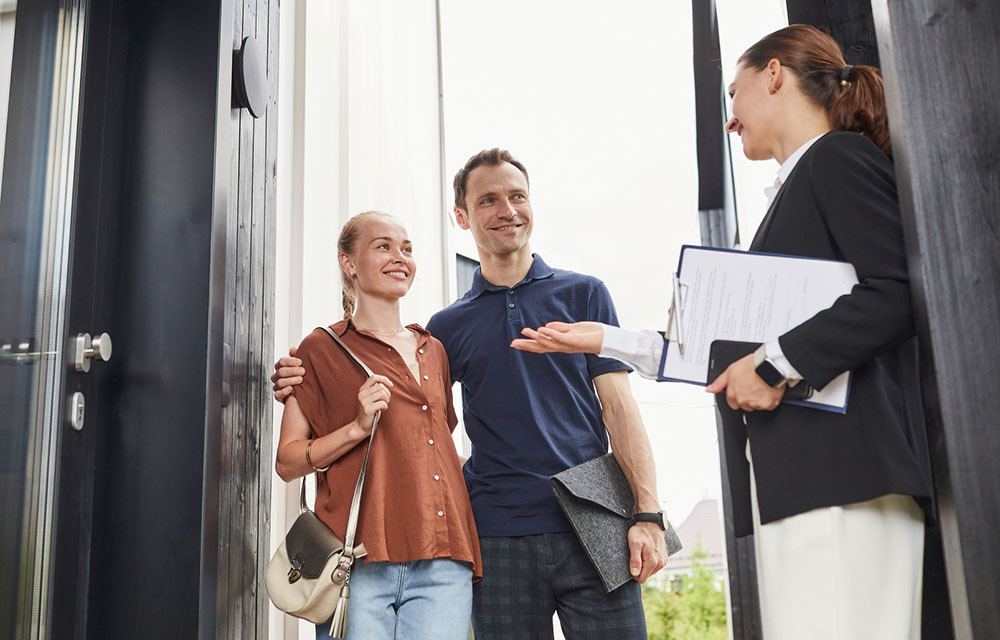 Real estate agent, holding a clipboard, meeting young couple at the doorstep of a house being shown.