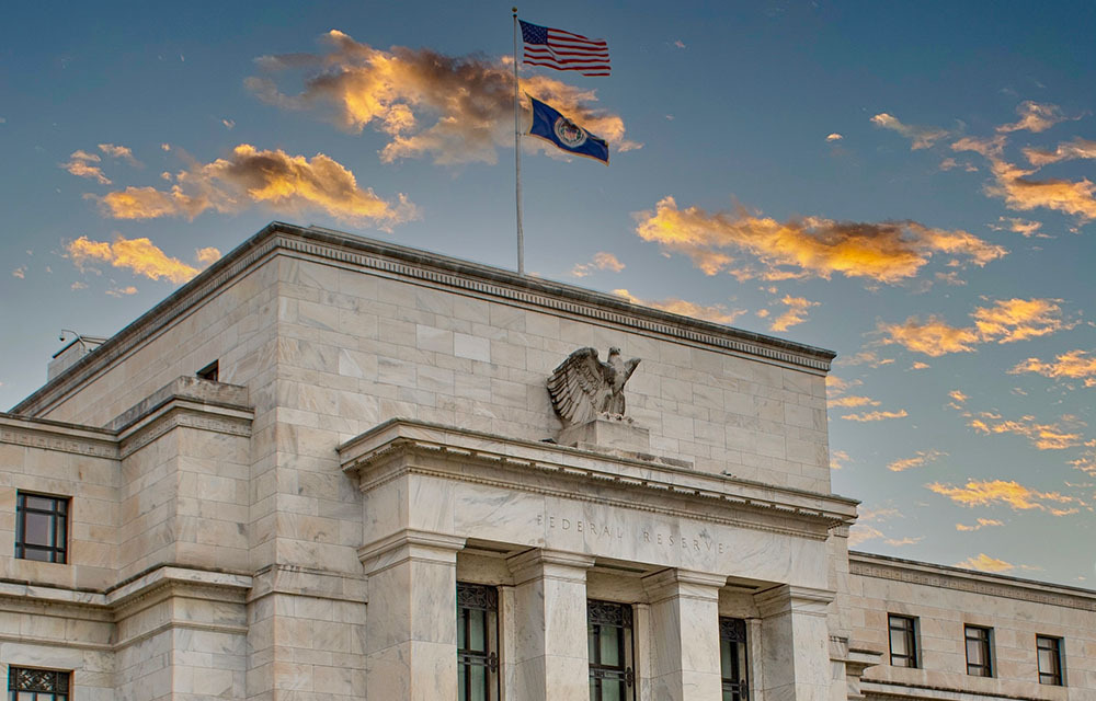An American flag and a flag with the Federal Reserve logo waving in the wind above the Board of Governors building.