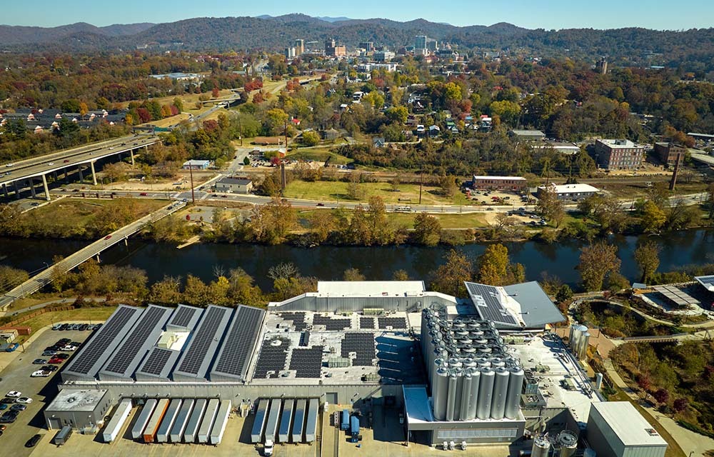 Aerial view of manufacturing plant with solar panels located in a rural, small town with many mountains in the distance.