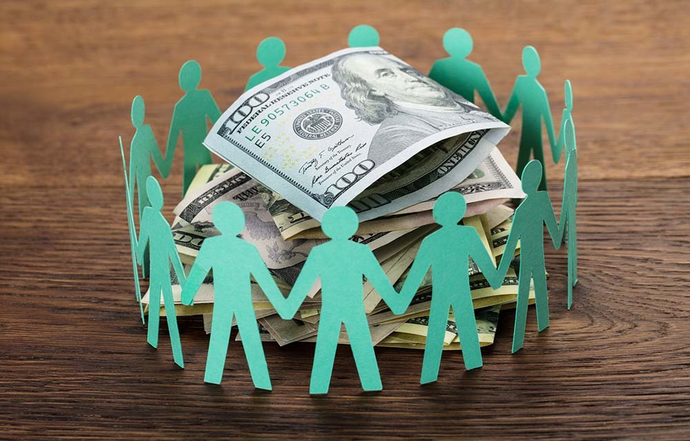 Paper cutout human figures holding hands around a stack of hundred-dollar bills.