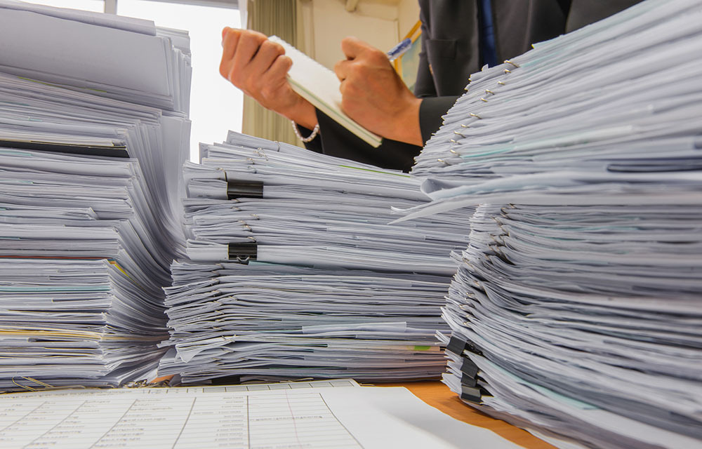 Stack of files on table as businessman writes on a notebook in the background.