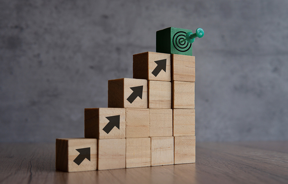 Wooden blocks stacked like a staircase with the blocks on the edge decorated with arrows. The block at the top has a target with a push pin in the bullseye.