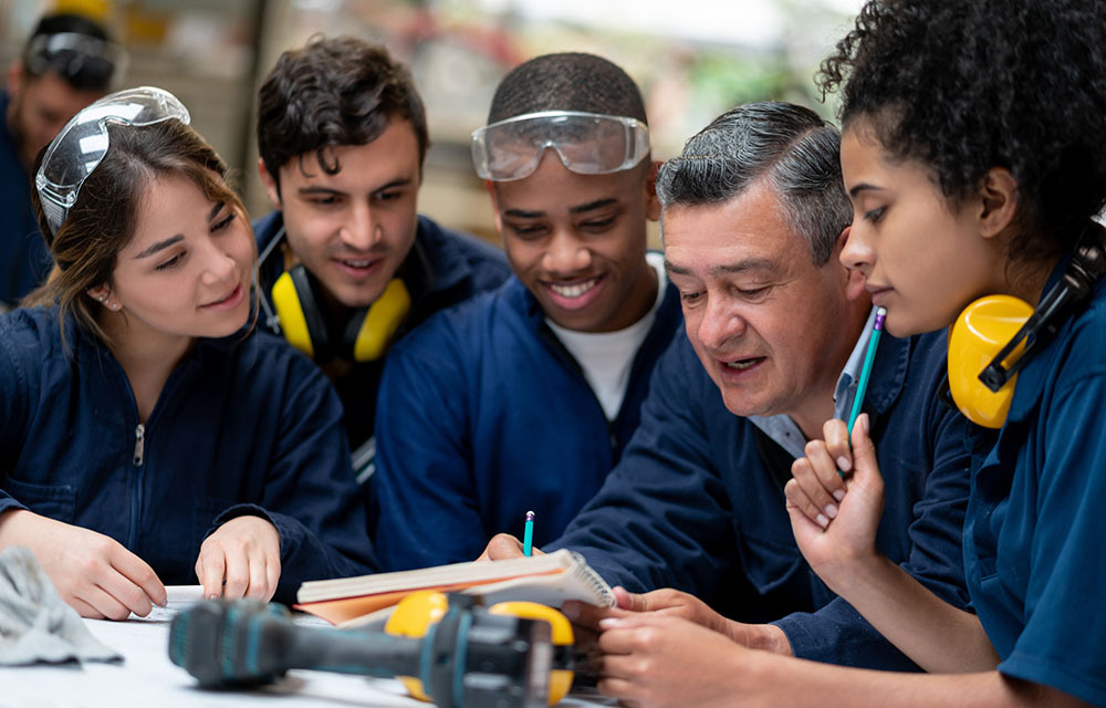 Apprentices dressed in blue overalls and safety equipment happily gathered around their teacher writing in a notebook.