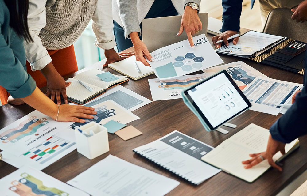 A group of business professionals brainstorm over a table with documents, charts, and infographics spread across.