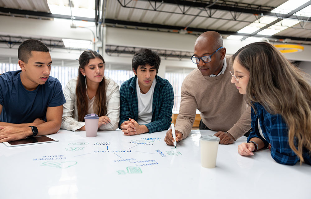 A professor and student gathered around a whiteboard on a table brainstorming ideas.