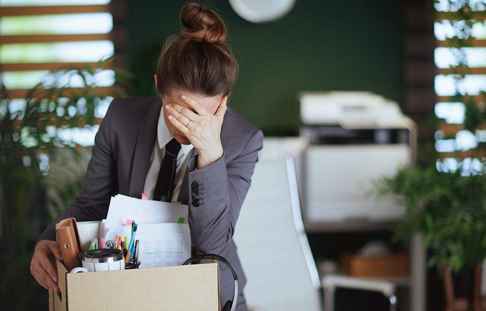 Distressed worker buries her face in her hand as she packs her office belongings into a box.