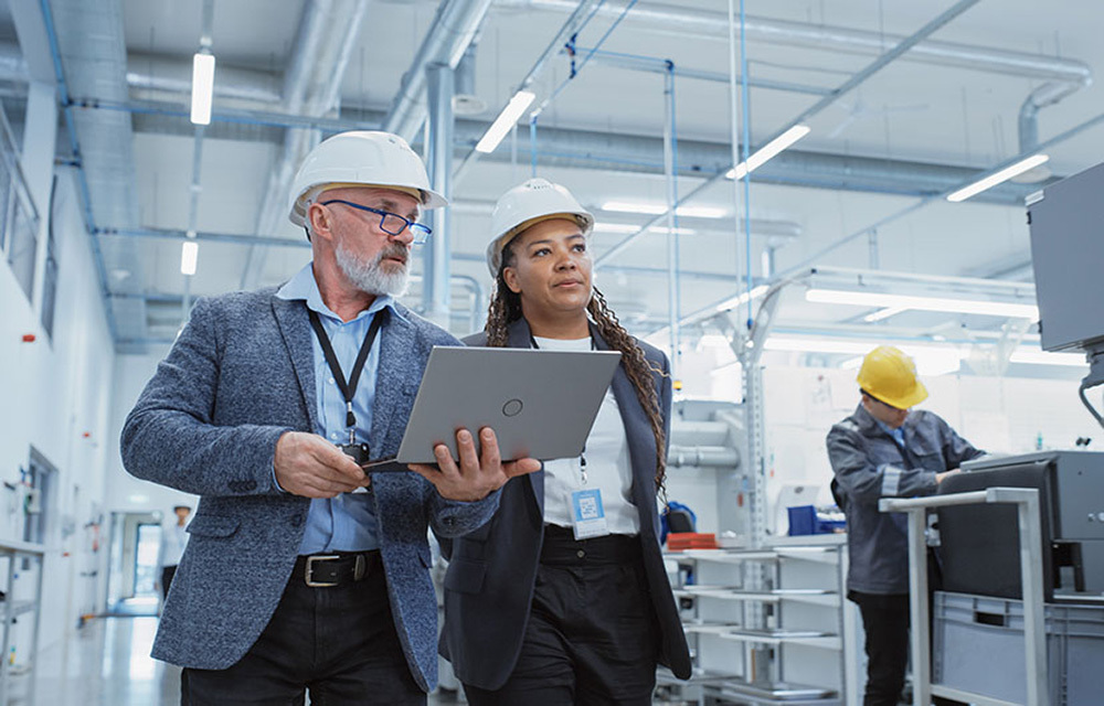 Two engineers in hardhats looking at laptop in hand while walking around factory.