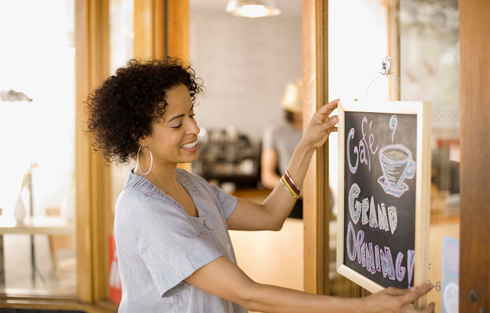 Woman hanging up a chalkboard that advertises a grand opening for her cafe.