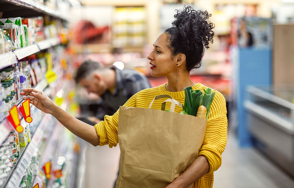A woman in a yellow sweater looking at products in a grocery store, carrying a paper bag with fresh vegetables peeking out.