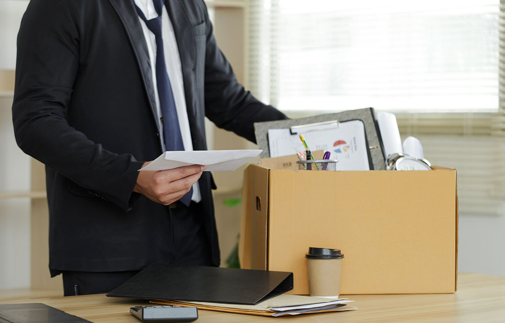 Man in suit holds his layoff letter as he gathers office materials into a box.