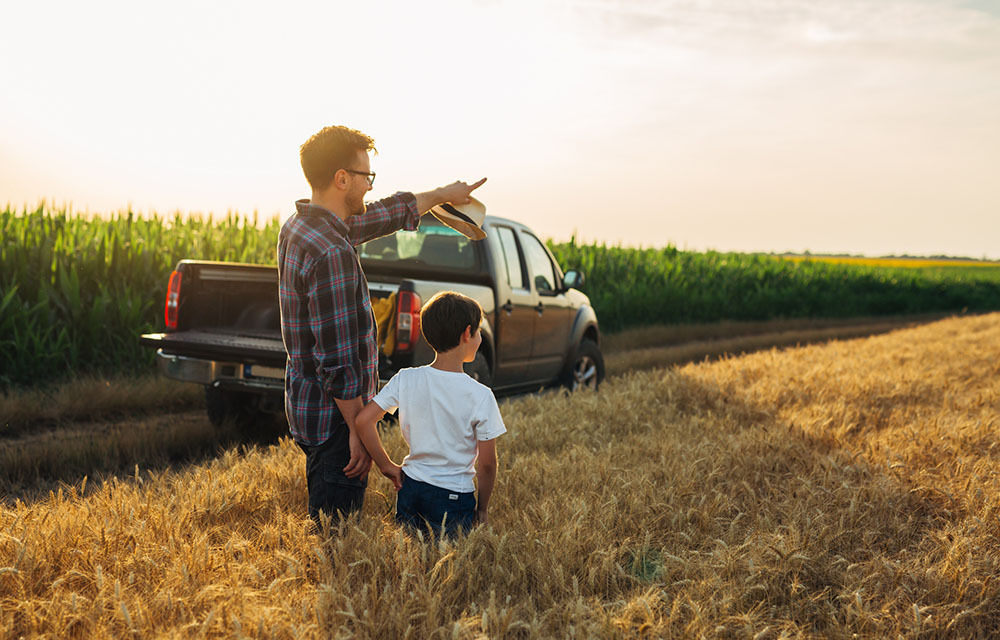 Father and son standing in a wheat field with a pickup truck in the background.