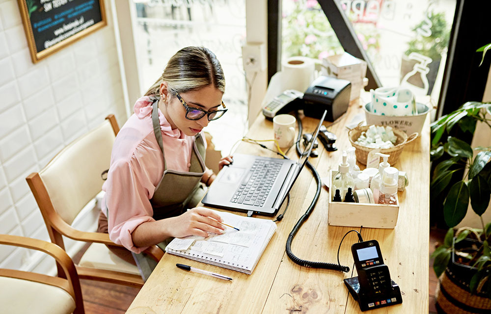 Small business owner, wearing an apron, sits at a desk as she adds up the day's sales in a notebook and enters the data into her laptop.