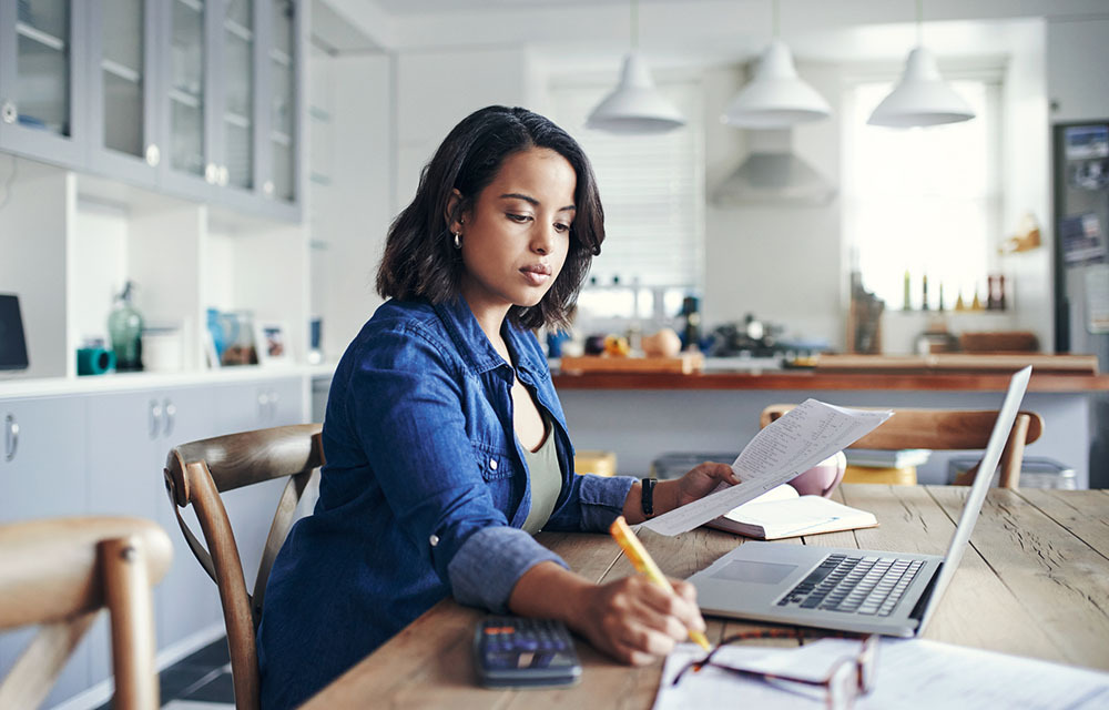 Woman working with a laptop, pencil, paper and calculator in kitchen at home