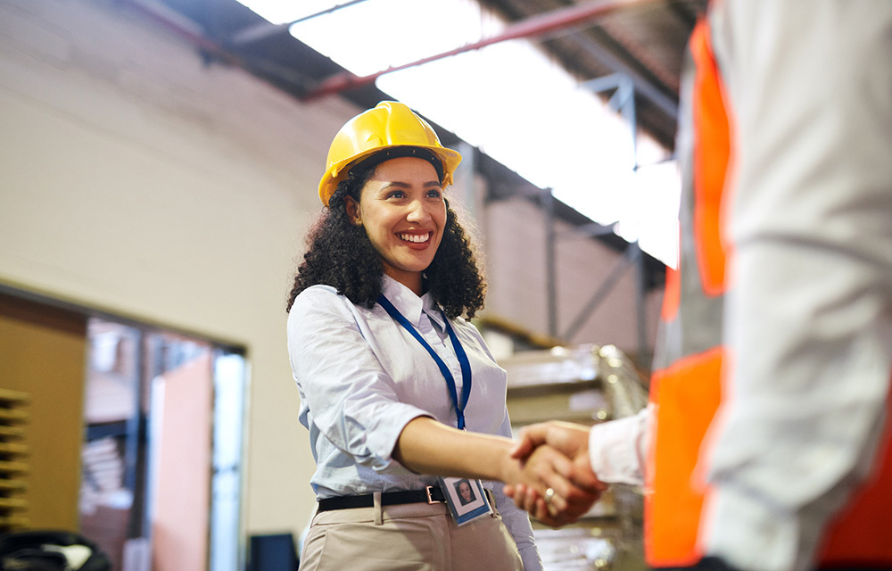 Woman wearing a hardhat smiling and shaking hands with worker in orange vest.