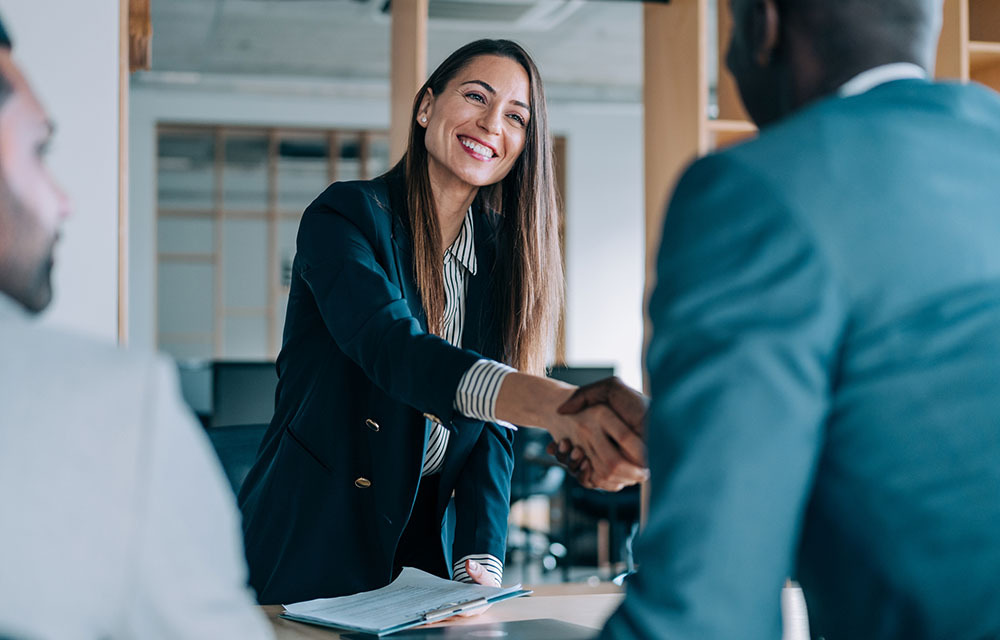 A woman and man in business attire smiling and shaking hands across a meeting table.