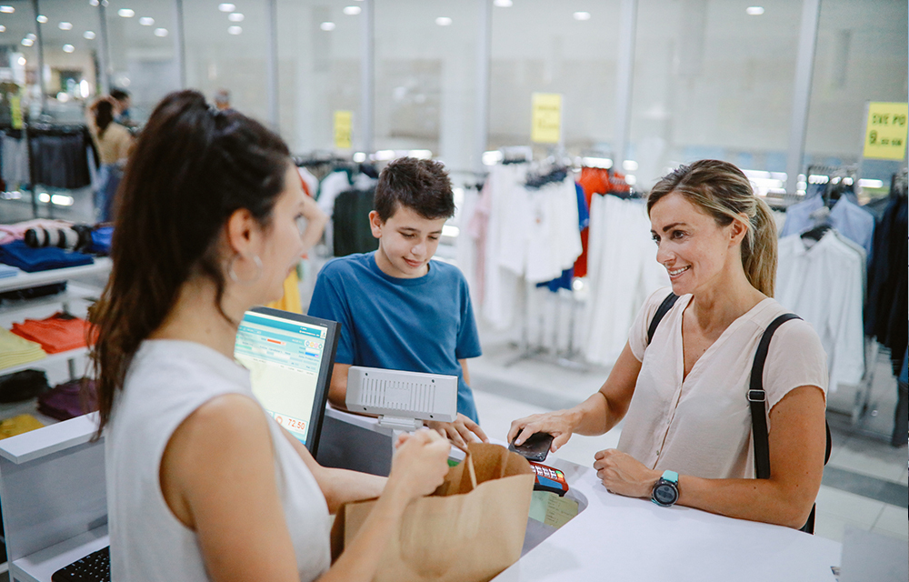 A woman buying her son new clothes in a store. She’s at the checkout, paying contactless with her smartphone.