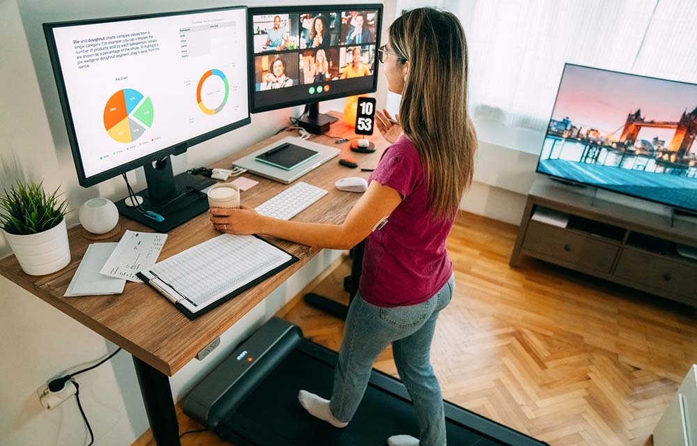 Woman at standing desk in home office talking on business video call while walking on treadmill.