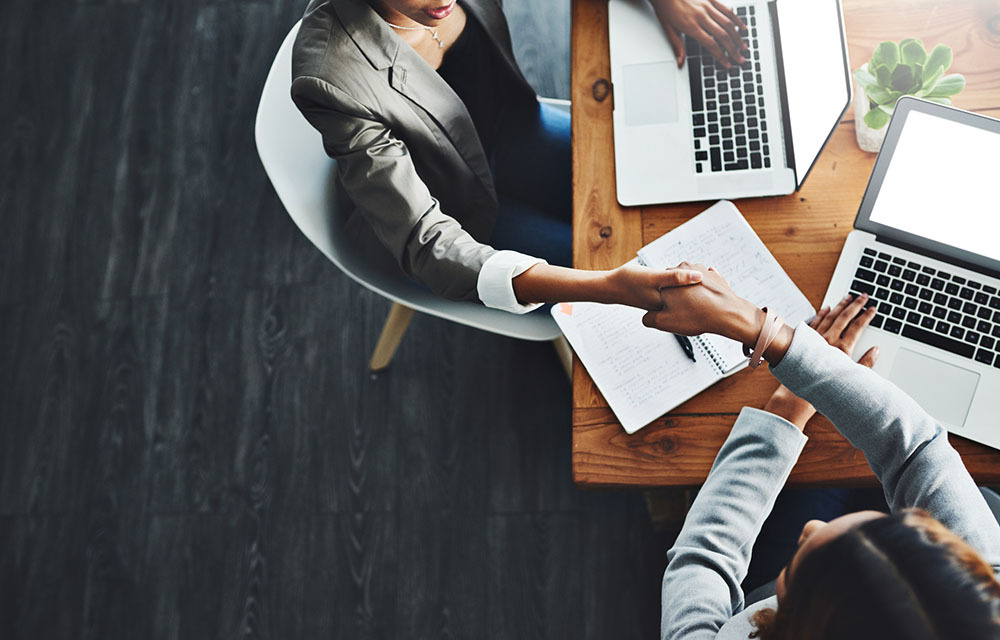 Two professional women shaking hands while seated at table with laptops in front of them.